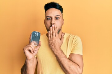 Young arab man holding canned food covering mouth with hand, shocked and afraid for mistake....