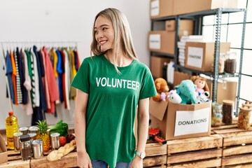 Asian young woman wearing volunteer t shirt at donations stand looking away to side with smile on face, natural expression. laughing confident.