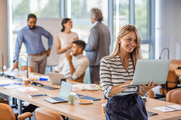 Woman manager holding laptop and standing in modern office. Businesswoman in casual wear holding laptop smiling and looking to camera while her colleagues communicating in the background