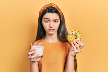 Young brunette girl eating donut and drinking glass of milk skeptic and nervous, frowning upset...