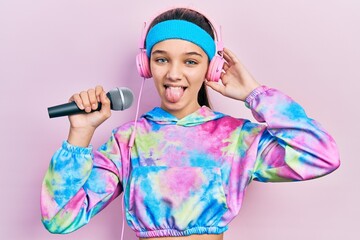 Young brunette girl singing song using microphone wearing sportswear sticking tongue out happy with...