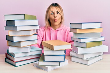 Young caucasian woman sitting on the table with books making fish face with lips, crazy and comical gesture. funny expression.