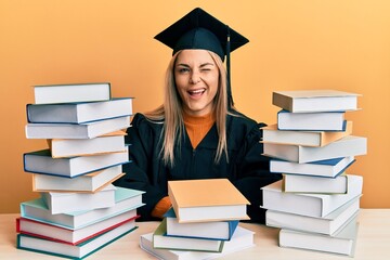 Young caucasian woman wearing graduation ceremony robe sitting on the table winking looking at the...