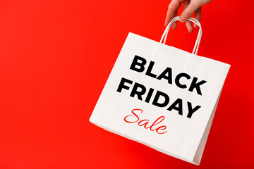 Female hand holding white shopping bag with Black Friday Sale text on red background. Sale, discount, shopping concept.
