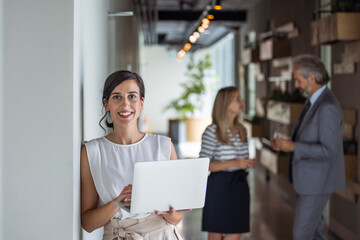 Portrait of a smiling confident young businesswoman holding laptop in hand looking at camera with...