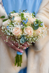 Winter bridal bouquet in hands. Beige roses, blue and gray flowers. Blue dress and beige fur coat on the bride. Wedding bouquet in pastel colors close-up. Flowers.