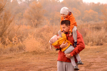 Father and two toddler children having fun on autumn day outdoors. Dad is carrying little girl in arms, boy is sitting on neck of father. Happy family spending good time together.