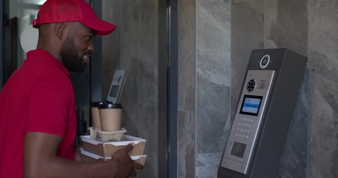 Courier african man doing fast food delivery, dials the house number in intercom. Bearded male in red uniformv came to customers, holding cardboard boxes and coffee in hands,very pleasant and nice