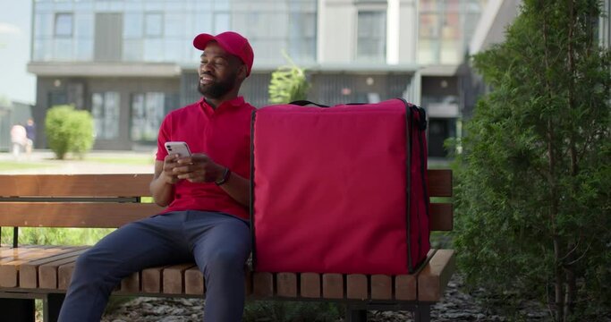 Afro american delivery male sit on bench talking with someone, with big red thermo bag with food. Black man in red uniform and cap looking at side on strange male, have conversation