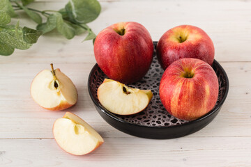 In a round black dish on white wooden table are three juicy beautiful ripe red apples of the early winter variety Gala. Next to the slices of the cut fruit. Top view