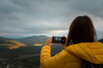 Photographer autumn landscape. A young woman tourist shoots a sunset in the mountains. Colorful autumn landscape, silhouette of a girl with a phone in her hands. Amateur photographer from behind