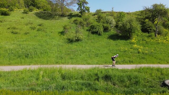 The speed skater in sport suit skating on a country road with beautiful green, hilly nature at background. Drone footage with parallel view of skater