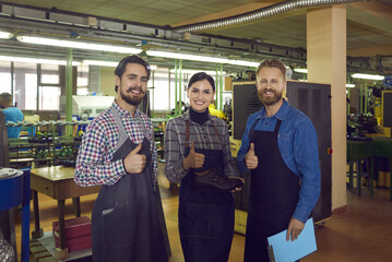 Portrait of three skilled happy male and female shoemakers at a shoe factory showing thumbs up. Two men and a woman standing in a large workshop room guarantee the good quality of their production.
