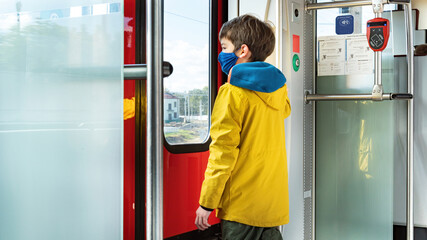 A child wearing a mask rides on public transport during the COVID 19 pandemic. A boy wearing a medical mask rides in an electric train looking out the window. Public transport privileges concept. - obrazy, fototapety, plakaty