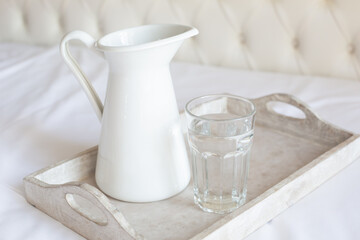 A white jug and a glass cup of water stand on a wooden tray on a white bed. Daily water intake. Retro style. 