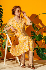 Fototapeta Fashionable redhead freckled woman wearing trendy summer midi dress, yellow square sunglasses, with classic leather bag, posing on yellow background. Full-length portrait obraz