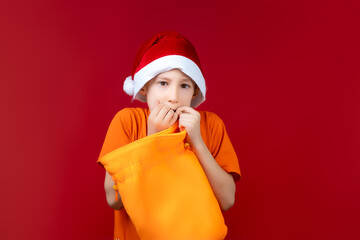 a boy in a Santa hat on a Christmas red background holds a yellow gift bag in his hands and looks fearfully into the camera
