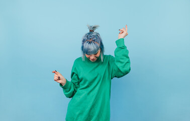 Positive woman with turquoise hair and in a green sweatshirt dancing on a blue background.
