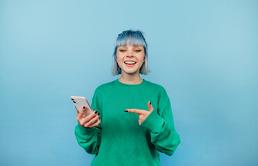 Positive bright lady in a green sweatshirt and colored hair stands on a blue background with a smartphone in his hands and looks at the camera with a smile on his face.