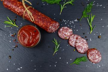 Salami sausage with ketchup and spices on black slate board. Top view