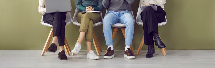 Legs of four job applicants sitting on chairs in hallway waiting in line for interview. Unknown candidates for jobs in casual clothes and shoes are sitting with documents and gadgets. Hiring concept. - obrazy, fototapety, plakaty