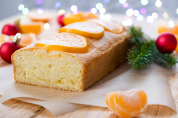 Loaf of gluten free mandarin cake with pieces of tangerine, fir branch, Christmas toys and lights on rustic wooden background. Citrus pie by classic recipe for winter holidays.