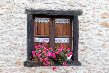 Fototapeta na wymiar Ventana rural con marco marón geranios y manualidades artesanales blancas, en una fachada de piedra, en el pueblo Calatañazor, Soria