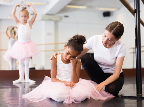 Young Female Classical Dance Teacher Comforting Tired And Crying Little Ballerina In Ballet Class