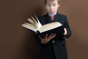 smart child 7-8 years old, kid in black school suit stands and reads thick tome, open paper book,...