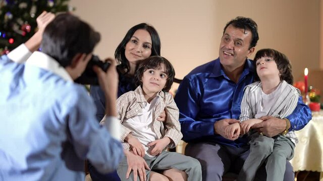Caucasian mother and Middle Eastern father with twin sons in living room on Christmas as teenage boy taking photo on camera. Concept of family celebration on New Year's eve