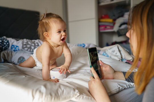 One small caucasian baby six months old naked in diapers on bed having fun smiling while her mother is taking photos using mobile phone real people domestic life concept family bonding leisure time