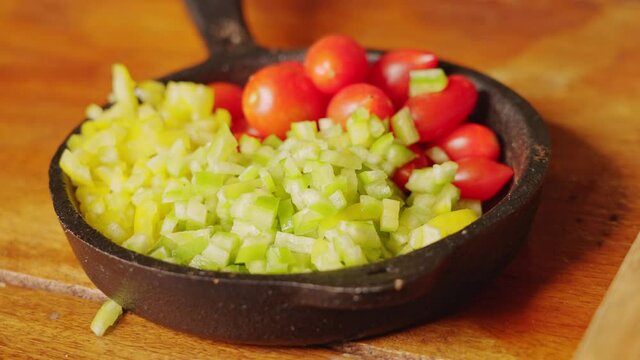 Handheld shot of a mini cast iron pan full of brunoise dice cut green capsicum and roma tomato for salad preparation.