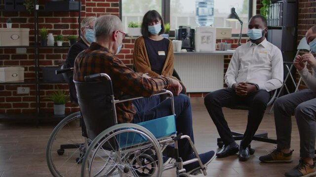 Senior man with disability attending aa group therapy session with people during pandemic. Person with face mask sitting in wheelchair and talking about alcohol addiction at meeting.