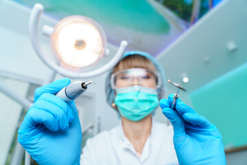 Dentist in a surgical mask, holding an angle mirror and a drill, ready to work. Woman dentist at work. Makes an examination and treatment of teeth for caries.