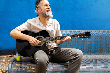 Mature adult caucasian man playing the guitar on a bench enjoying a sunny day.