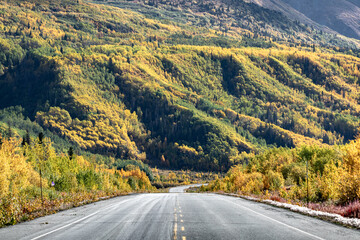 Driving through fall covered landscape colored road in northern Canada on the Alaska Highway with golden mountains and stunning boreal forest surrounding the road. 