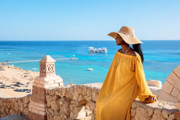 Happy African American woman in yellow dress and sun hat enjoys view of coast of Red Sea on natural background. Panoramic views of blue sea with yachts and coastline, Sharm El Sheikh, Egypt.  - Powered by Adobe