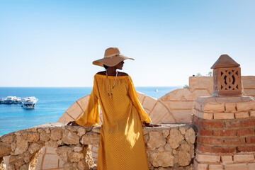 Happy African American woman in yellow dress and sun hat enjoys view of coast of Red Sea on natural background. Panoramic views of blue sea with yachts and coastline, Sharm El Sheikh, Egypt.  - Powered by Adobe