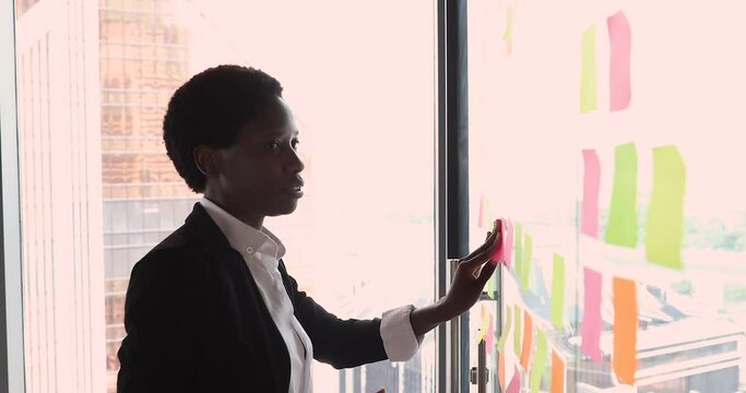 Serious African businesslady preparing boardroom for negotiations corporate meeting attaching sticky notes on glass wall. Start up ideas creation, strategy development, make schedule or agenda concept