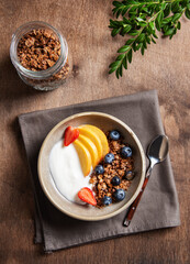 Fresh natural yogurt with homemade granola, peach, strawberry and blueberry in a bowl on a napkin and dark wooden background. The concept of a healthy and nutritious breakfast. Diet food. Top view