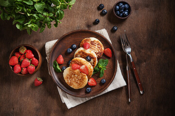 Cottage cheese pancakes or syrniki with blueberries, strawberries and fresh mint on a dark wooden background. The concept of a home-made healthy breakfast. Top view and copy space image