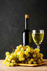 a glass glass and a bottle of white wine and a brush of white grapes on a cutting board on a black background