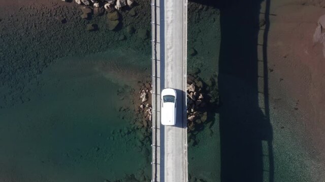 White car parked in middle of bridge with calm blue water on sunny day, aerial