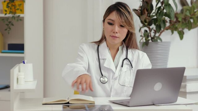 Doctor telehealth remote service. Asian female traumatologist in white robe writes email and looks at wrist x-ray photo at desk in hospital office spbi 4k video
