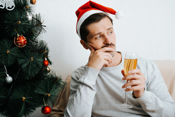 Mental health, loneliness, sad depressive mood for Christmas celebration. Upset lonely young man in santa hat with glass of champagne sitting alone at home near decorated xmas tree and looking away
