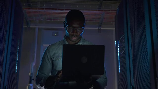 American African male engineer working with laptop and walking along big data office spbas. Closeup view of young man holds computer in his hands and looks at screen, inspects equipment racks and