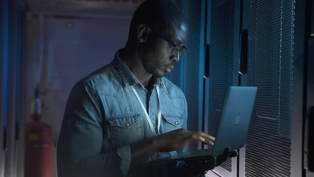 Young male engineer using laptop and inspecting equipment while standing in big data center spbas. Closeup view of American African man holds computer and checks hardware racks, types report and does