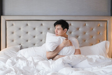 Young male sitting on bed in the morning after getting up. Handsome young man resting sitting in his bed. Young man in white t shirt feeling comfortable and sitting on bed in morning.