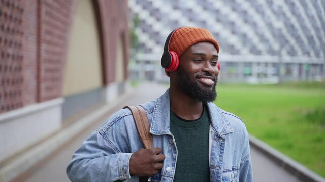 Modern devices to listen music. Happy young black guy hipster in denim jacket and warm orange hat smiles enjoying music sounds with earphones on city street spbas