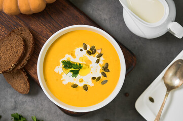 Pumpkin soup with parsley, cream and pumpkin seeds in a white bowl, top view. Layout on gray background with ingredients and bread